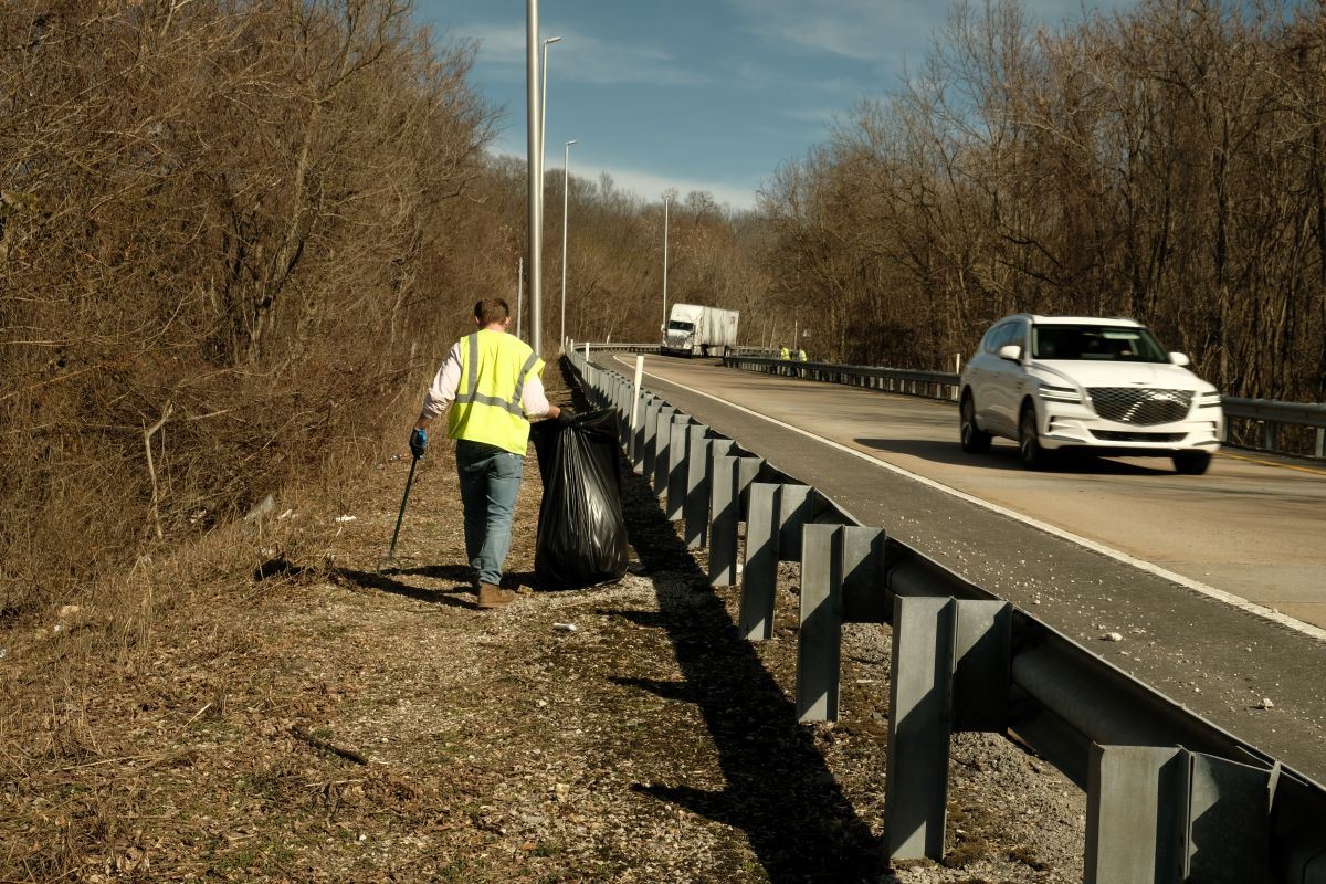 Maintenance workers in yellow vests picking up litter and debris from along roadside.